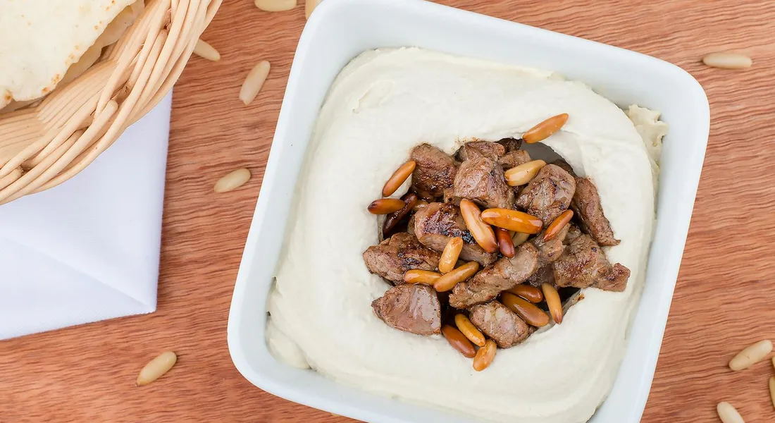 Close-up of a white bowl containing a creamy dip topped with pine nuts and small chunks of meat