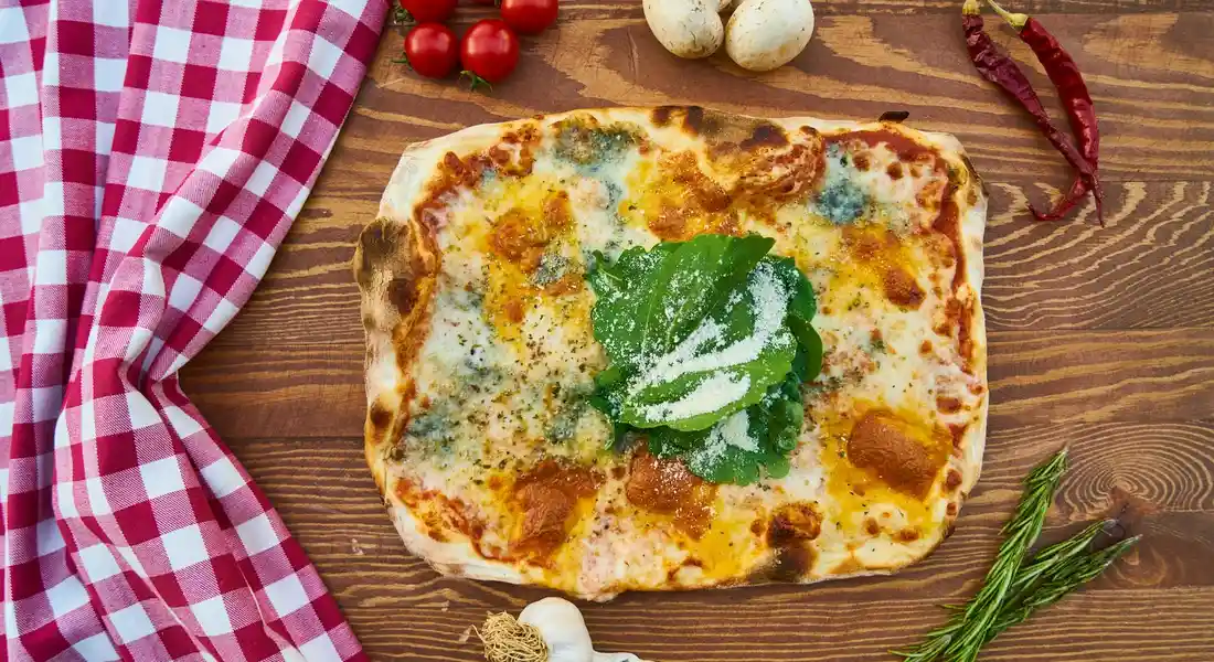 Cheesy pizza on a wooden table with a red-and-white checkered napkin, garlic, tomatoes, and herbs nearby.