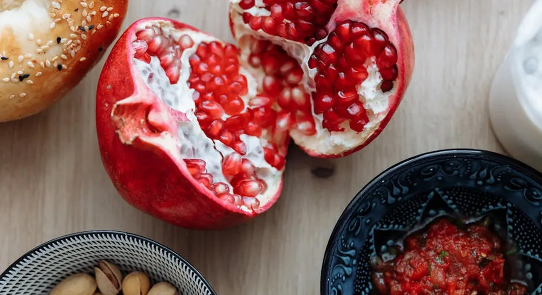 Close-up of a split pomegranate with bright red arils on a wooden surface, with small bowls nearby.