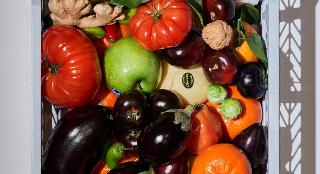 Assorted fresh vegetables in a crate, including eggplants, tomatoes, peppers, and apples.