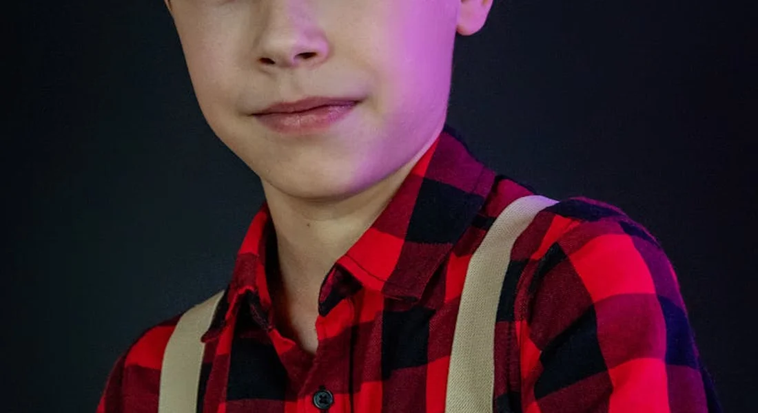 Close-up of a person wearing a red and black plaid shirt with beige suspenders, with purple lighting on one side of the face