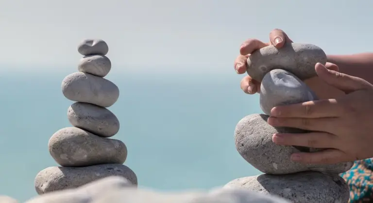 Close-up of hands stacking smooth stones on a beach with the ocean in the background.