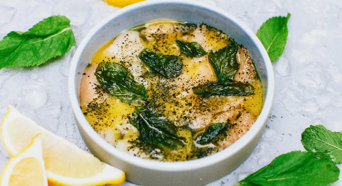 A white bowl containing a dish garnished with fresh mint leaves and cracked pepper, with lemon wedges visible at the edge.