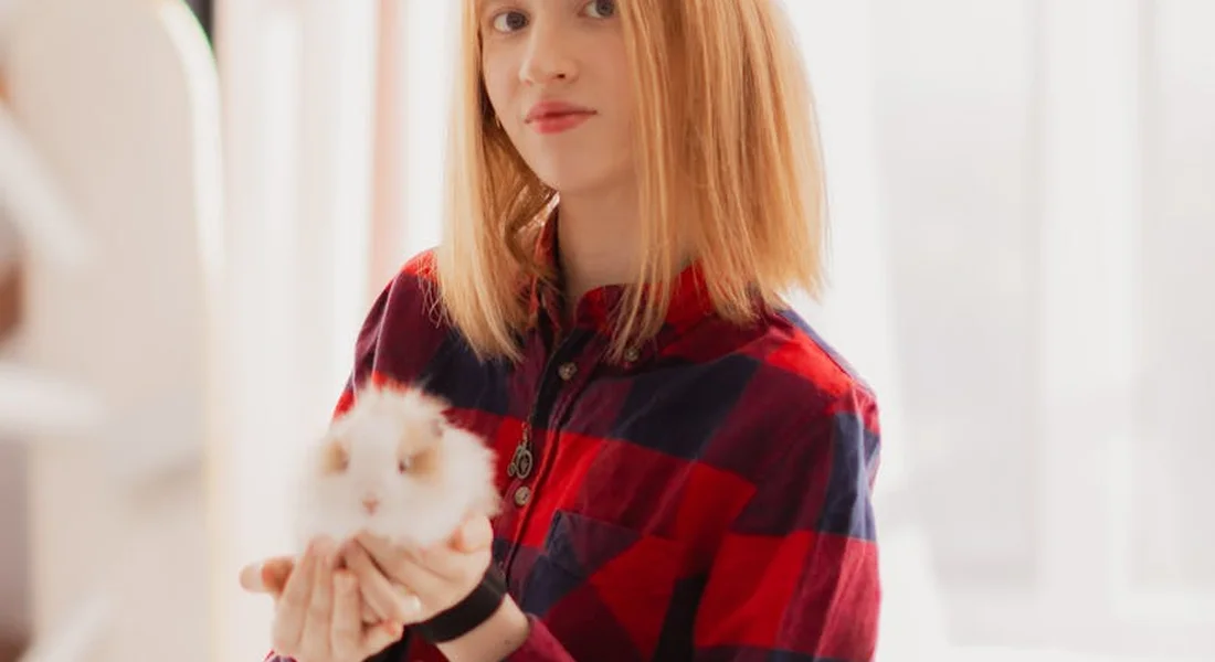 A person with blonde hair wearing a red and black plaid shirt gently holds a small white guinea pig indoors.