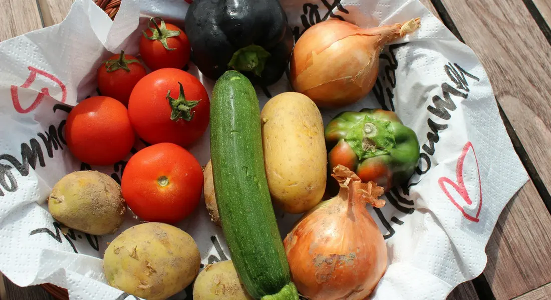 Assorted fresh vegetables—tomatoes, zucchini, potatoes, onions, and peppers—arranged on a paper-lined basket on a wooden surface.