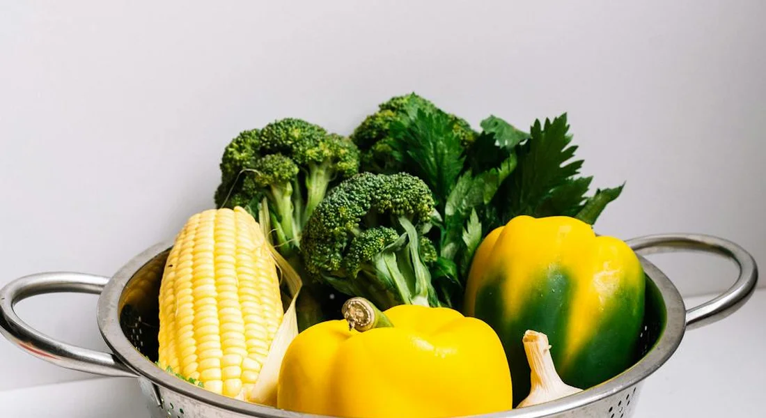 Fresh vegetables (broccoli, yellow bell pepper, corn, and leafy greens) in a metal colander, illustrating natural vitamin C sources for guinea pigs.