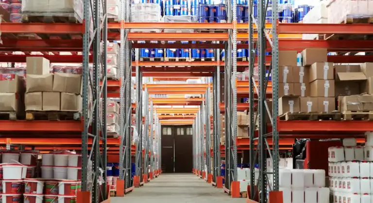 Industrial warehouse with tall orange shelving units, pallets, and boxed goods arranged in an aisle.