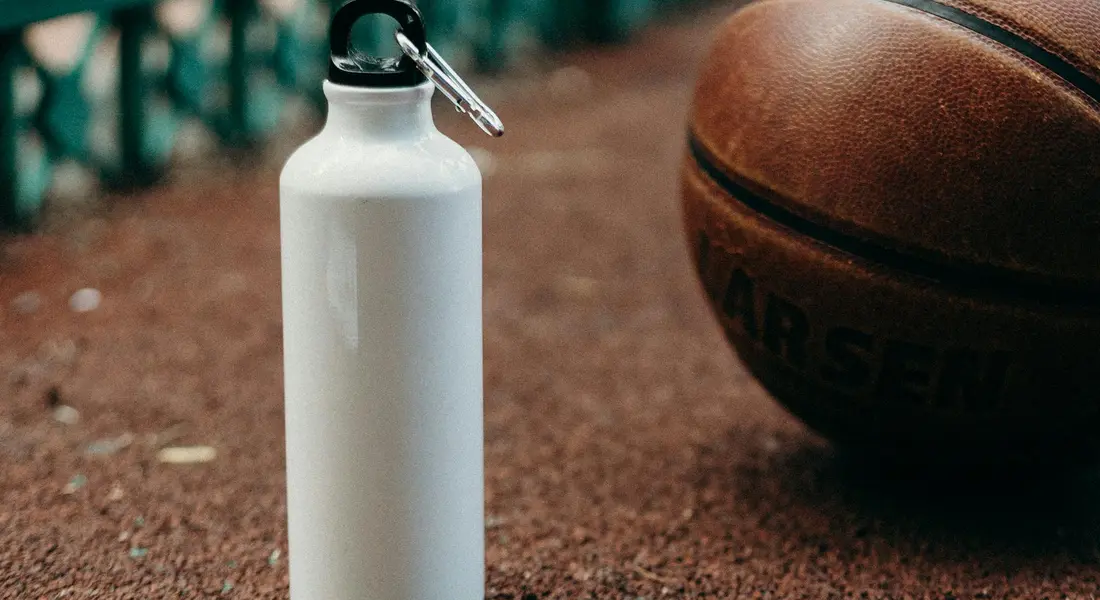 White metal water bottle with a drinking spout on a textured outdoor surface next to a basketball.