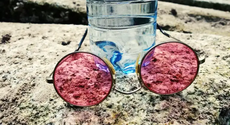 Clear plastic water bottle on a rough rock surface with round pink sunglasses in front.