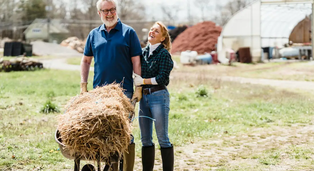 Two adults on a farm standing beside a wheelbarrow filled with hay, with a barn and farming equipment in the background.