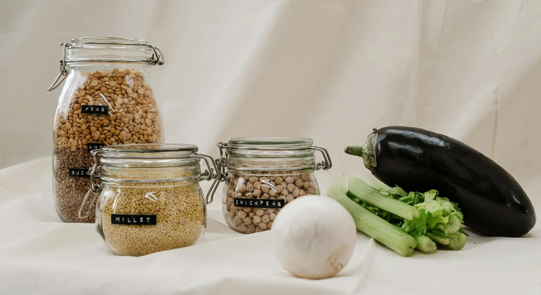 Glass jars with airtight lids containing dry goods on a pale surface, with a white onion, celery, and a dark eggplant nearby—suggesting organized storage ideas for keeping guinea pig pellets fresh.
